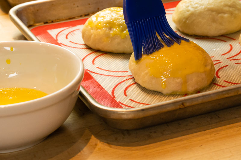 Silicone basting brush coating pretzel buns with egg wash in preparation for baking.