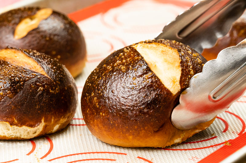 Metal tongs grabbing a fresh-baked pretzel bun