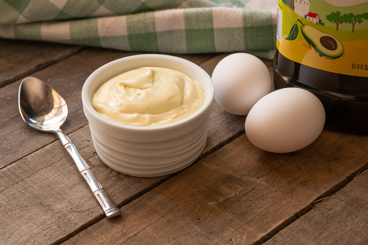 A bowl with homemade mayonnaise, a spoon, two eggs, a bottle of avocado oil, and a checkered towel