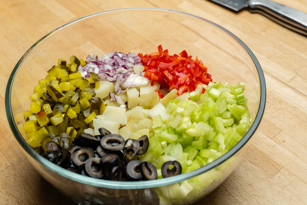 Diced pickles, potatoes, red bell pepper, red onion, olives, and celery in a clear glass bowl.