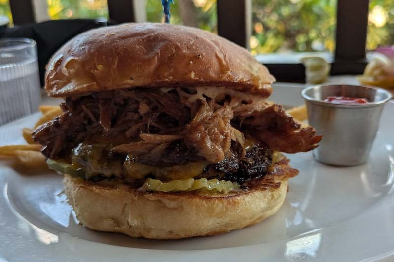A cheeseburger with pulled pork, bbq sauce, garlic aioli, pickles, on a toasted bun. Along with a side of french fries and a container of ketchup.